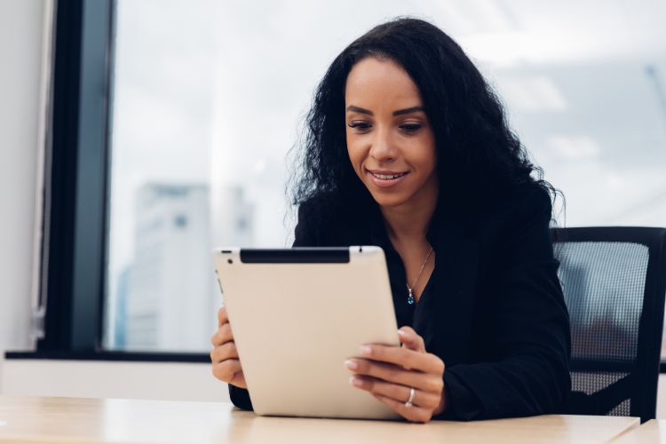 Uma mulher está sentada em uma cadeira, segurando um tablet e olhando para a tela com atenção. Ela está em um ambiente de escritório, com uma janela ao fundo que mostra uma vista urbana desfocada.