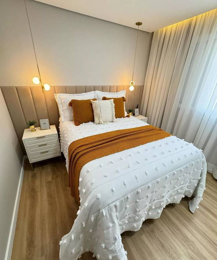 Um quarto de casal decorado com tons neutros e detalhes em laranja. A cama possui uma colcha branca texturizada e uma manta laranja, além de almofadas brancas e laranjas. Ao lado da cama, há dois criados-mudos brancos com luminárias pendentes. As cortinas claras e o piso de madeira completam o ambiente aconchegante.