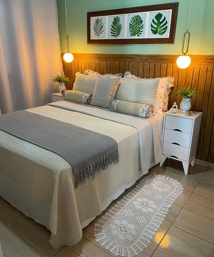 Quarto de casal com parede verde e cabeceira de madeira. A cama tem colcha bege, manta cinza e almofadas decorativas. Há luminárias pendentes e um quadro com folhas verdes na parede.