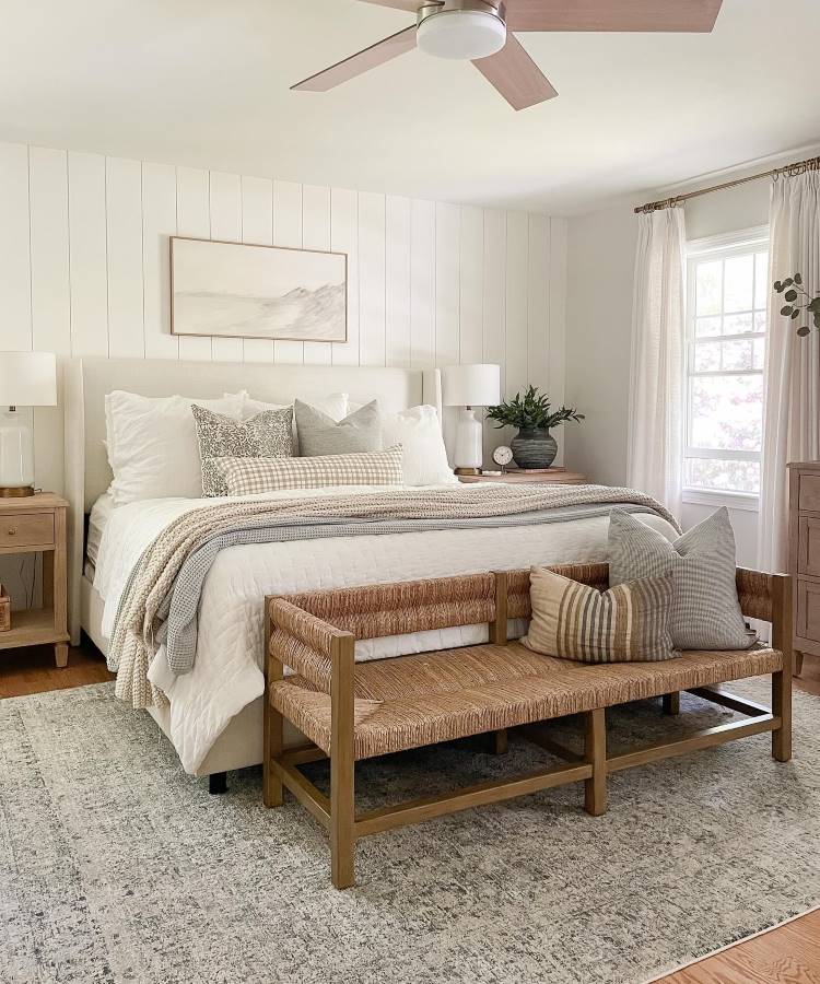 Quarto de casal com cama branca, almofadas em tons neutros e um banco de madeira aos pés da cama. A decoração inclui luminárias de mesa e uma grande janela com cortinas claras