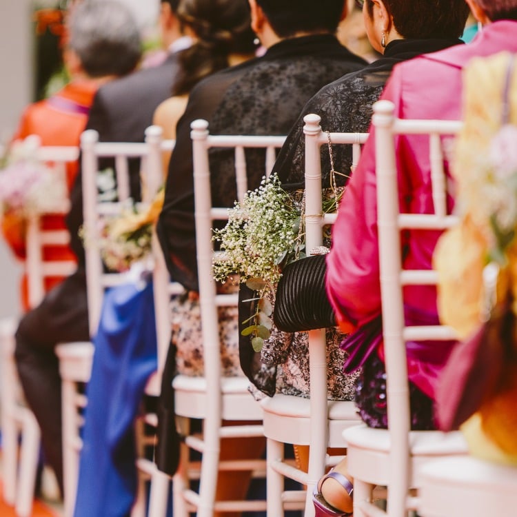 A imagem mostra uma fileira de cadeiras brancas em um evento, provavelmente um casamento. As cadeiras estão decoradas com pequenos arranjos de flores brancas. As pessoas sentadas estão vestidas de forma elegante, com roupas coloridas e formais. A cena transmite um ambiente festivo e organizado, típico de uma cerimônia de casamento.