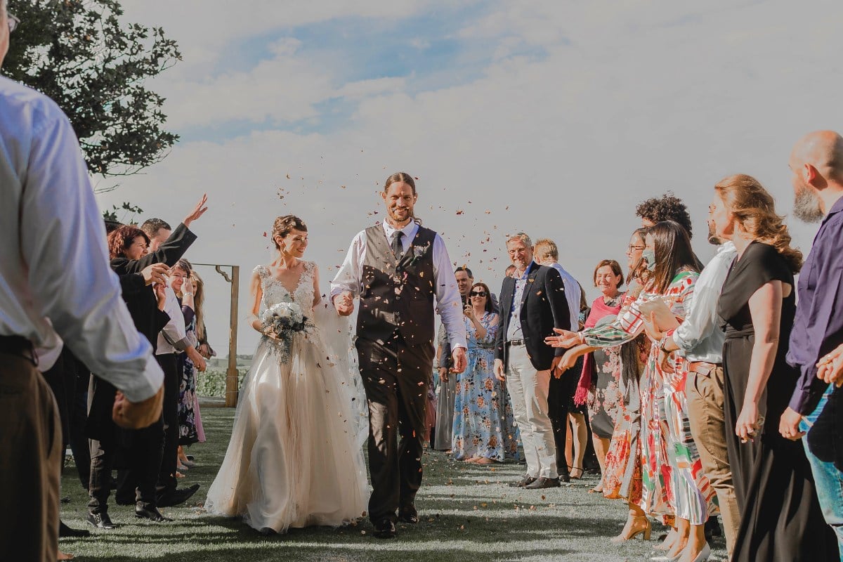 A imagem mostra um casal recém-casado caminhando por um corredor ao ar livre, cercado por convidados. A noiva está vestida com um vestido de noiva branco e segura um buquê, enquanto o noivo usa um colete e gravata. Os convidados, dispostos em duas fileiras, parecem estar jogando pétalas ou confetes no casal, celebrando o momento. O ambiente é ensolarado, com céu azul ao fundo, criando uma atmosfera alegre e festiva.