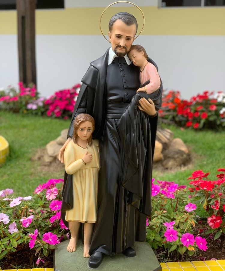 A imagem mostra uma estátua de São Vicente de Paulo, um santo católico, vestindo uma túnica preta. Ele segura um bebê adormecido em seus braços e uma criança está ao seu lado, olhando para ele. Um halo dourado circunda sua cabeça. Ao fundo, há flores coloridas em um jardim.