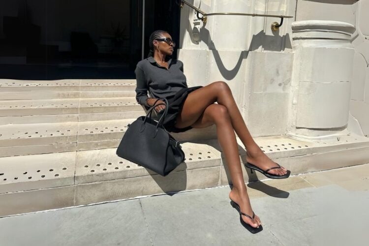 Foto de mulher negra sentada em degraus usando look preto e sandália de dedo, uma das tendências de sapato para o verão 2025