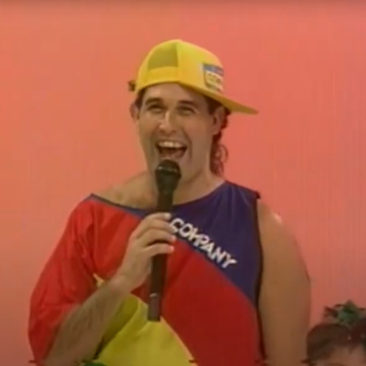 Sérgio Mallandro nos anos 80, durante a apresentação de um programa infantil. Ele está usando uma camiseta colorida e um boné amarelo virado para o lado, segurando um microfone enquanto sorri amplamente. A imagem captura sua energia contagiante e estilo descontraído, característicos de sua carreira na televisão.