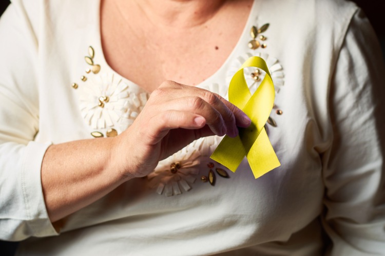 A imagem mostra uma pessoa vestindo uma blusa clara com detalhes florais em relevo. Ela está segurando um laço amarelo, que simboliza a conscientização sobre a prevenção do suicídio (setembro amarelo). O laço está preso à blusa, destacando-se contra o tecido. A cena é iluminada de forma suave, destacando a importância do símbolo.