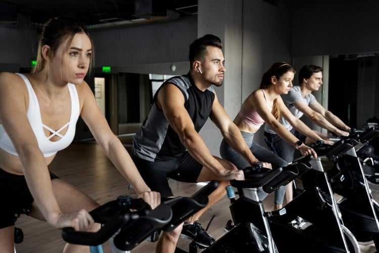 Foto de duas mulheres e dois homens em academia praticando spinning. A primeira mulher usa top branco e shorts preto, o primeiro homem usa bermuda preta e regata cinza e preta, a segunda mulher usa top rosa e calça cinza e o segundo homem usa blusa cinza e bermuda preta 