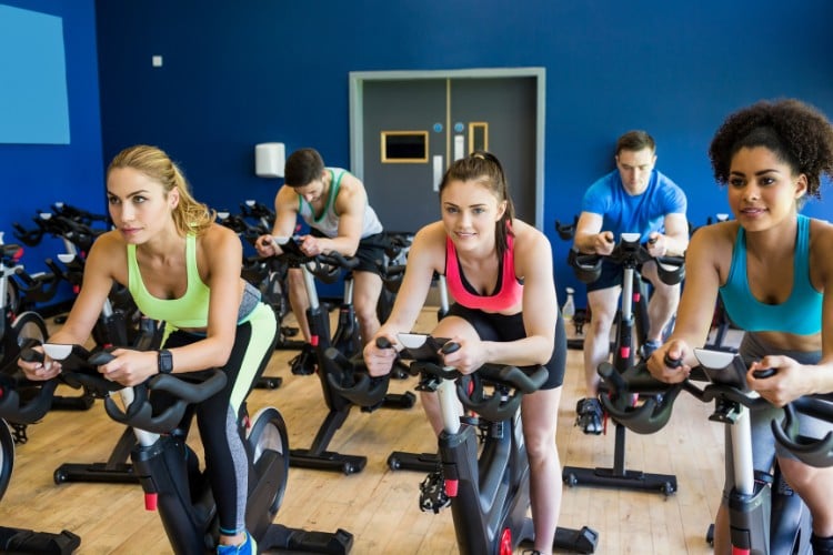 4 pessoas de pele clara e uma de pele negra praticando spinning em academia de parede azul