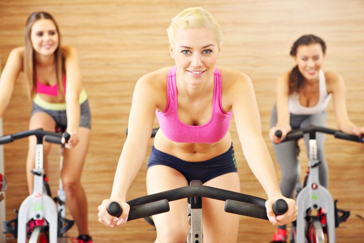 3 mulheres de pele clara fazendo bicicleta, sendo a da esquerda com top rosa e shorts cinza; a do meio com top rosa e shorts azul marinho e a da direita usando top branco e calça cinza