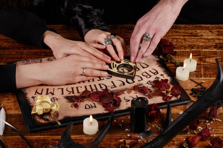 A imagem mostra um tabuleiro Ouija sobre uma mesa de madeira. Três mãos descansam sobre a planchette, que está posicionada no centro do tabuleiro. As mãos usam anéis, incluindo um com uma caveira. Ao redor do tabuleiro, há pétalas de rosa secas, velas acesas, cristais e um chifre escuro. A cena tem uma atmosfera mística e esotérica.