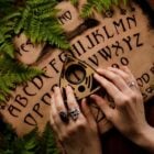 A imagem mostra um tabuleiro Ouija cercado por folhas verdes, possivelmente samambaias, em um fundo de madeira. Duas mãos estão posicionadas sobre a planchette, que é triangular e possui um círculo no centro. As letras do alfabeto e números estão dispostos no tabuleiro, junto com as palavras "sim" e "não". As mãos usam anéis, incluindo um com uma caveira. A cena tem uma atmosfera mística e esotérica.