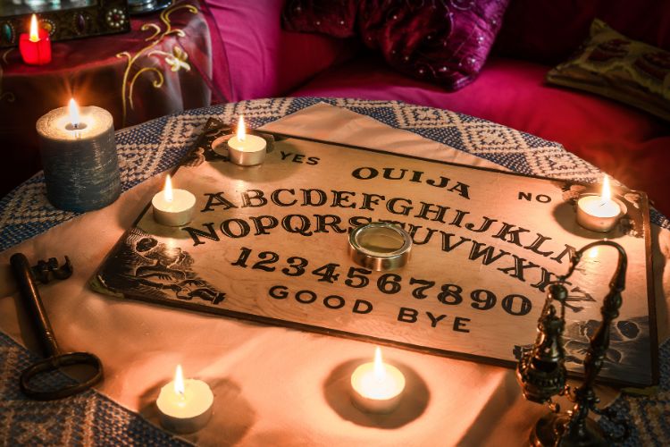 A imagem mostra um tabuleiro Ouija colocado sobre uma mesa coberta por um tecido decorativo. O tabuleiro exibe o alfabeto, números de 0 a 9, e as palavras "sim", "não" e "adeus". Ao redor do tabuleiro, há várias velas acesas, criando uma atmosfera mística e intimista. Também há uma chave antiga e um incensário próximo, adicionando um elemento esotérico à cena. O ambiente parece acolhedor, com almofadas coloridas ao fundo.