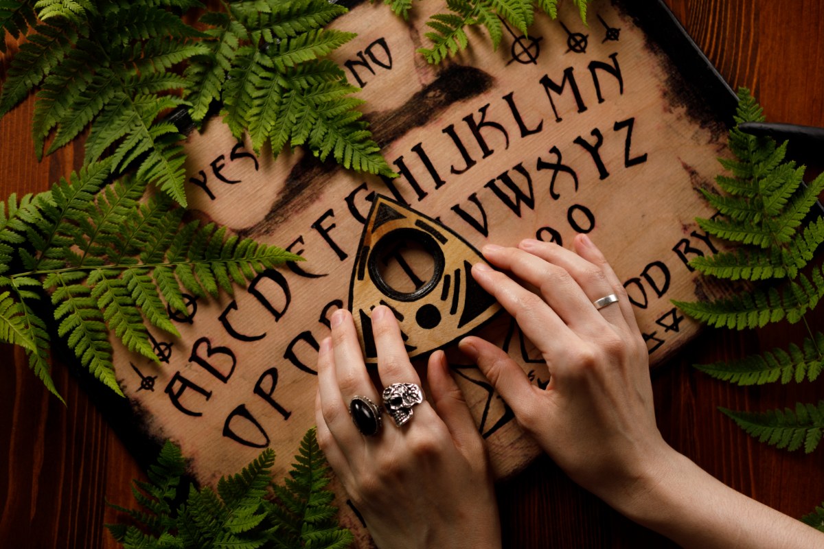 A imagem mostra um tabuleiro Ouija cercado por folhas verdes, possivelmente samambaias, em um fundo de madeira. Duas mãos estão posicionadas sobre a planchette, que é triangular e possui um círculo no centro. As letras do alfabeto e números estão dispostos no tabuleiro, junto com as palavras "sim" e "não". As mãos usam anéis, incluindo um com uma caveira. A cena tem uma atmosfera mística e esotérica.