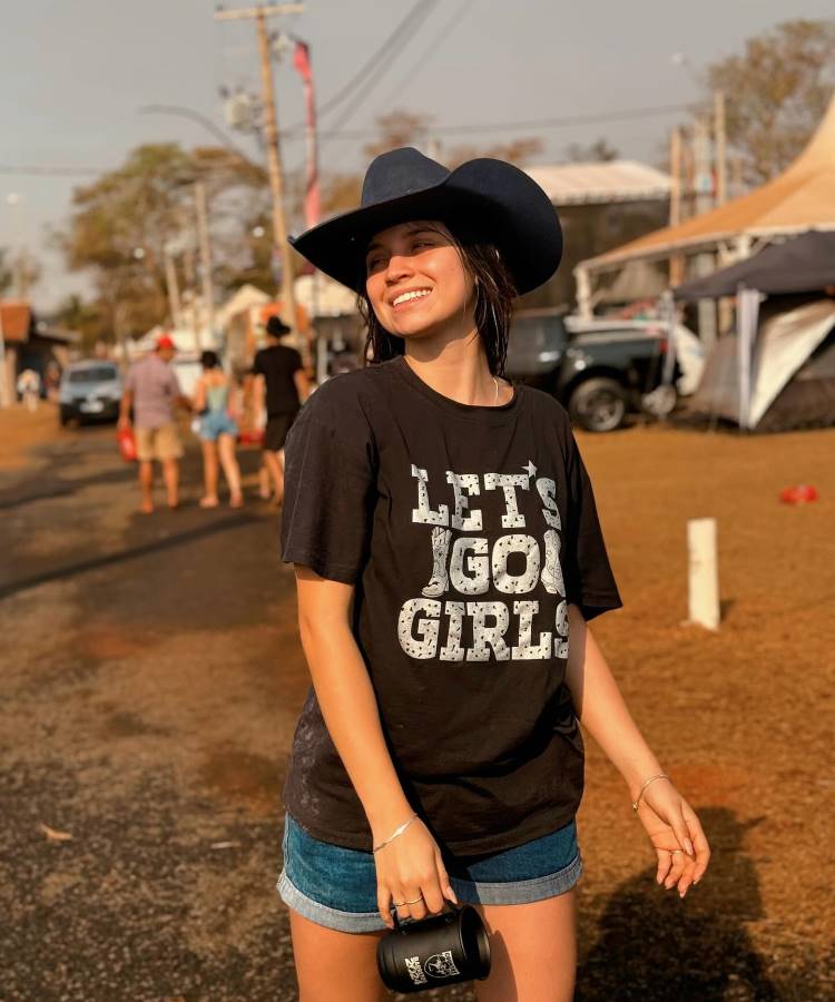 A imagem mostra uma pessoa usando um chapéu e uma camiseta preta com a frase "LET'S GO GIRLS". O tipo de decote da camiseta é redondo, característico por ser simples e circular, sem muitos detalhes.