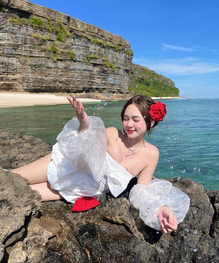 Uma mulher deitada em rochas à beira-mar, usando um vestido branco com mangas bufantes e uma flor vermelha no cabelo. Ao fundo, há um penhasco rochoso e o mar azul sob um céu claro.