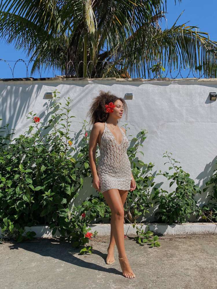 Julia Rodrigues usando vestido de crochê branco com plantas ao fundo e flor presa na orelha 