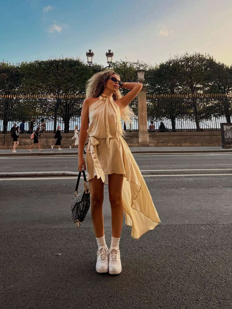 Julia Rodrigues usando vestido amarelo manteiga assimétrico, com tênis e bolsa preta