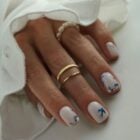 Mão usando anéis dourados e de pérolas com unhas curtas decoradas em tons de azul e branco