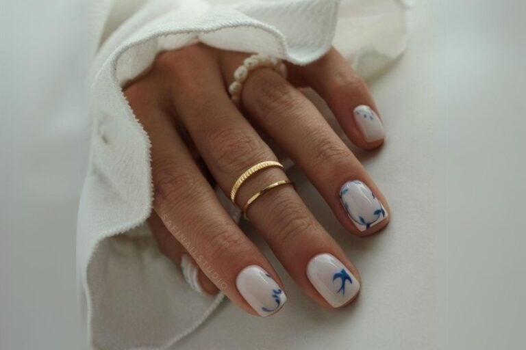 Mão usando anéis dourados e de pérolas com unhas curtas decoradas em tons de azul e branco