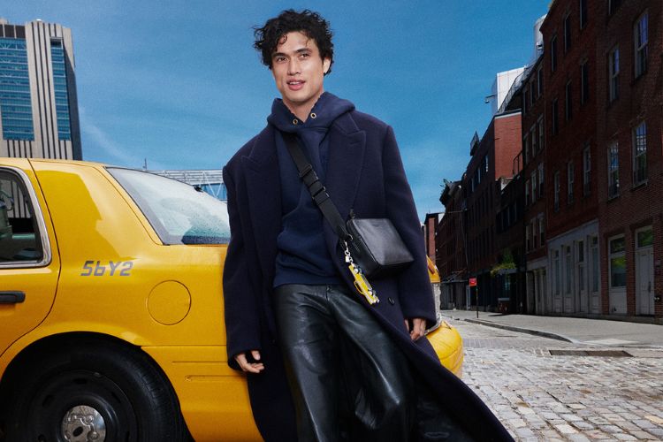 Charles Melton em frente a um táxi amarelo usando moletom, calça, sobretudo e bolsa, tudo em preto