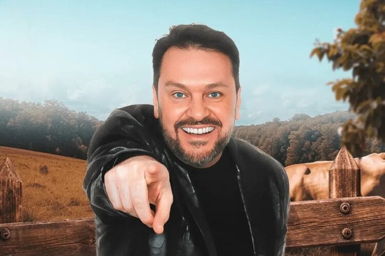 A imagem mostra Albert Bressan sorrindo e apontando para a câmera. Ele veste uma jaqueta preta. Ao fundo, há uma paisagem rural com uma cerca de madeira, campos e árvores sob um céu azul.