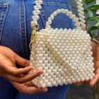 A imagem mostra uma pessoa segurando uma pequena bolsa feita inteiramente de pérolas brancas. A bolsa tem uma alça e detalhes em metal dourado. A pessoa veste uma calça jeans e usa pulseiras prateadas em ambos os pulsos. Ao fundo, há uma planta e o ambiente parece estar ao ar livre.