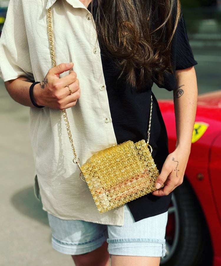 A imagem mostra uma pessoa usando uma camisa de duas cores, bege e preta, combinada com shorts jeans claros. Ela segura uma bolsa de ombro feita de esferas douradas com alça de corrente dourada. No pulso, há uma pulseira preta e um anel delicado. Ao fundo, há um carro vermelho parcialmente visível.