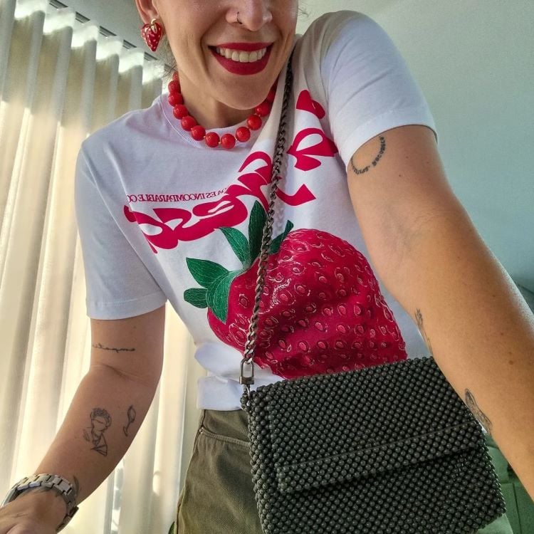 A imagem mostra uma pessoa sorridente vestindo uma camiseta branca com uma estampa grande de morango vermelho. Ela usa acessórios vermelhos, como brincos de coração e um colar de contas. A pessoa carrega uma bolsa de miçangas pretas com alça de corrente. Há várias tatuagens visíveis em seus braços, e o fundo exibe uma cortina clara.