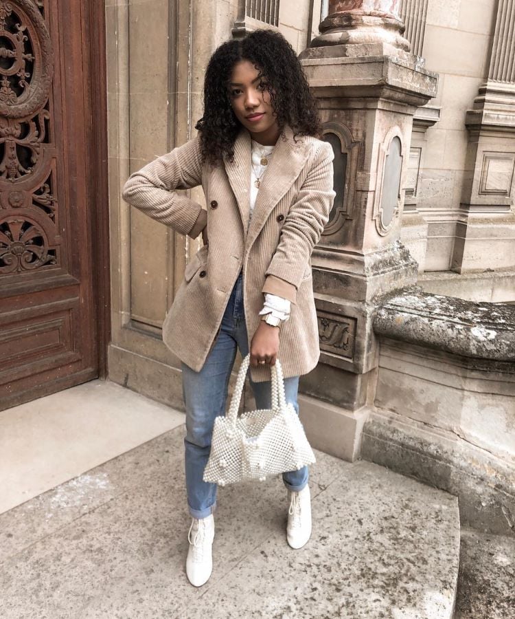 A imagem mostra uma mulher posando em frente a um edifício de arquitetura clássica. Ela veste um casaco bege de veludo cotelê, jeans azul claro e botas brancas. A mulher segura uma bolsa de pérolas brancas, e usa várias pulseiras no pulso esquerdo. Seu cabelo é cacheado e solto, e ela olha diretamente para a câmera.