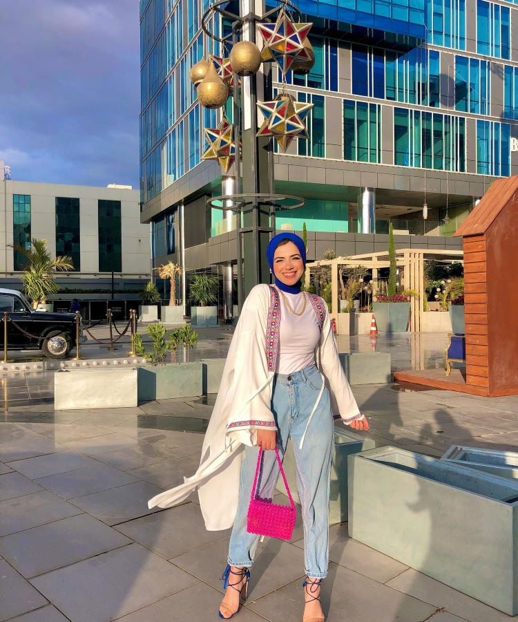 A imagem mostra uma mulher sorridente usando um hijab azul, blusa branca, calça jeans clara e um casaco longo com detalhes coloridos. Ela segura uma bolsa rosa vibrante e usa sandálias de salto azuis. Ao fundo, há um edifício moderno de vidro e metal, com decorações de estrelas douradas. O ambiente é urbano e iluminado pela luz do sol.