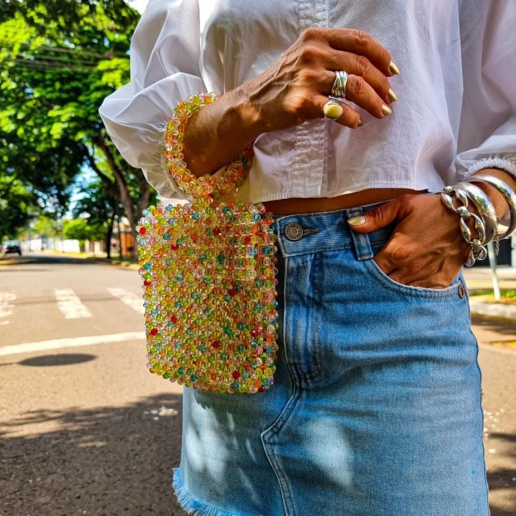 A imagem mostra uma pessoa usando uma blusa branca e uma saia jeans segurando uma bolsa feita de contas coloridas. A mão da pessoa exibe anéis grandes e pulseiras prateadas. O cenário ao fundo é uma rua arborizada, com luz natural, sugerindo um ambiente ao ar livre em um dia ensolarado. A bolsa tem alça curta.