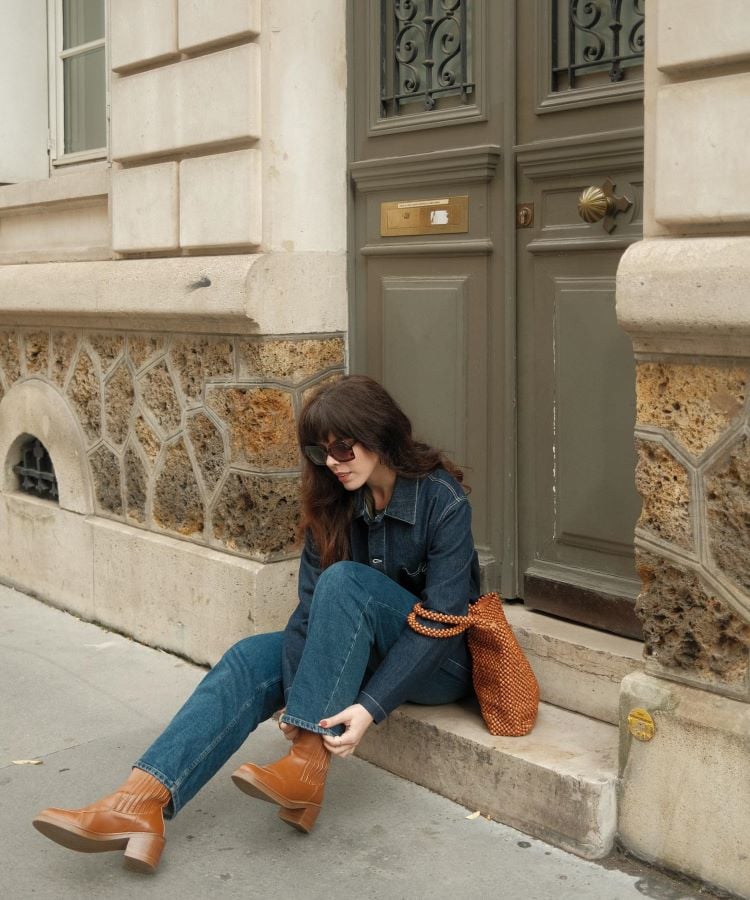 A imagem mostra uma mulher sentada em uma calçada, próxima a uma porta de madeira verde com detalhes em ferro. Ela veste jaqueta e calça jeans, botas marrons e usa óculos escuros. Ao lado, há uma bolsa de crochê laranja. O cenário é uma rua com paredes de pedra e arquitetura clássica. A mulher parece ajustar suas botas.