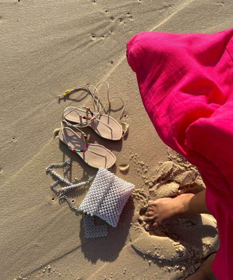 A imagem mostra uma pessoa de pé na areia, vestindo uma saia rosa vibrante. Ao lado, há um par de sandálias claras com detalhes coloridos e uma bolsa de pérolas brancas com alça de corrente. A areia tem marcas de pegadas e está iluminada pelo sol, criando uma atmosfera de praia relaxante e ensolarada.