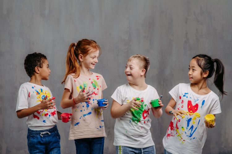 A imagem mostra quatro crianças sorrindo e interagindo enquanto seguram potes de tinta colorida. Elas estão vestindo camisetas brancas manchadas de tinta, indicando que participaram de uma atividade de pintura. As crianças parecem felizes e estão em pé contra um fundo cinza, destacando a vivacidade das cores em suas roupas. A cena transmite um sentimento de diversão e criatividade.