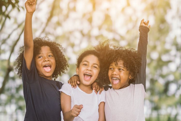 A imagem mostra três crianças sorrindo e demonstrando alegria. Elas estão juntas, com os braços levantados, em um ambiente ao ar livre. A expressão de felicidade delas transmite um momento de diversão e amizade. O fundo desfocado sugere um ambiente natural, possivelmente um parque, com luz suave e agradável.