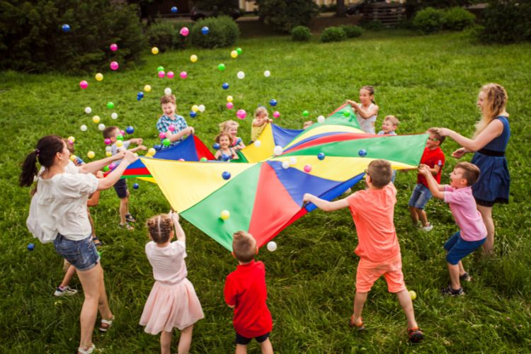 A imagem mostra um grupo de crianças brincando ao ar livre em um gramado. Elas estão segurando um grande tecido colorido, com seções em vermelho, amarelo, azul e verde. As crianças estão espalhadas ao redor do tecido, balançando-o para lançar várias bolas coloridas ao ar. A cena transmite alegria e diversão, com as crianças interagindo em um ambiente de brincadeira coletiva. Ao fundo, há vegetação, sugerindo que a atividade ocorre em um parque ou área verde.