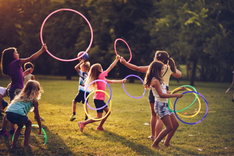 A imagem mostra um grupo de crianças brincando em um gramado ao ar livre, cada uma segurando um bambolê colorido. Elas estão em movimento, girando os bambolês ao redor de seus corpos. O ambiente é iluminado pela luz do sol, que cria uma atmosfera alegre e vibrante. Ao fundo, há árvores que sugerem um parque ou área verde. As crianças parecem estar se divertindo e participando de uma atividade coletiva.