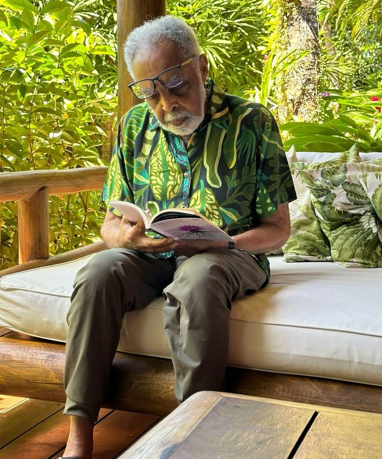 A imagem mostra um homem de óculos sentado em um sofá ao ar livre, em um ambiente cercado por vegetação. Ele está lendo um livro e veste uma camisa com estampa de folhas e calças cáqui. O cenário sugere um local tranquilo e natural, com almofadas decorativas ao fundo.