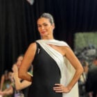 Na imagem, Carol Ribeiro está usando um vestido elegante e moderno, de um ombro só, com um design assimétrico. O vestido é predominantemente preto com um detalhe branco que cruza o corpo, criando um contraste sofisticado. Ela complementa o look com cabelos presos de forma elegante e brincos discretos, destacando sua elegância natural no evento. O fundo parece ser de um tapete vermelho, com pessoas e câmeras ao redor, sugerindo um ambiente de premiação.