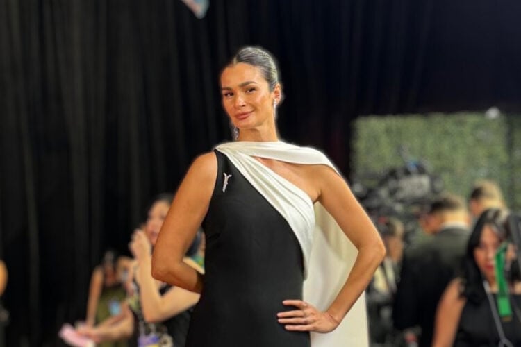 Na imagem, Carol Ribeiro está usando um vestido elegante e moderno, de um ombro só, com um design assimétrico. O vestido é predominantemente preto com um detalhe branco que cruza o corpo, criando um contraste sofisticado. Ela complementa o look com cabelos presos de forma elegante e brincos discretos, destacando sua elegância natural no evento. O fundo parece ser de um tapete vermelho, com pessoas e câmeras ao redor, sugerindo um ambiente de premiação.