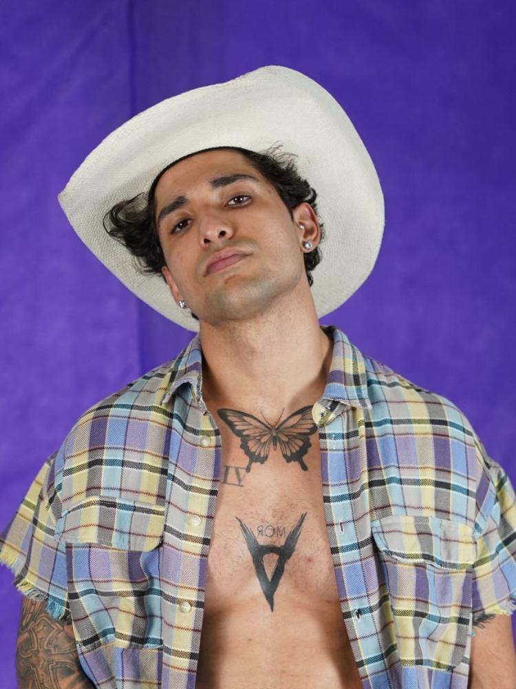 Cauê Fantin usando camisa xadrez azul e amarela aberta, revelando seu tronco com tatuagens, e chapéu de cowboy bege 