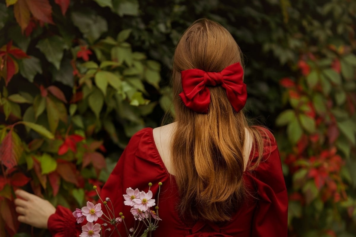 A imagem mostra uma mulher de costas, com cabelos longos adornados por um grande laço vermelho. Ela veste um vestido vermelho, combinando com o laço. Ao seu redor, há uma folhagem verde com toques de vermelho, criando um contraste harmonioso. Na mão, ela segura delicadas flores rosa, adicionando um toque suave à cena.