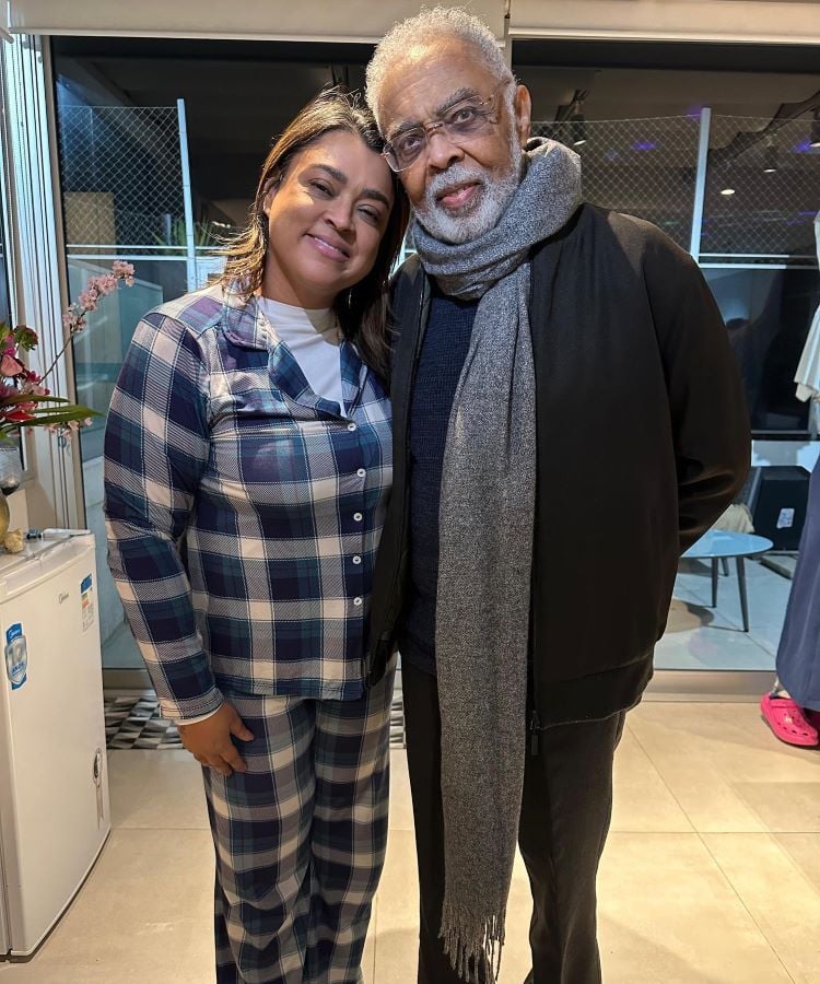 A imagem mostra duas pessoas abraçadas. Preta Gil está usando um pijama xadrez azul e branco, enquanto Gilberto Gil à direita está vestindo um casaco escuro e cachecol cinza. Elas estão em um ambiente interno, com uma porta de vidro ao fundo e algumas plantas decorativas.