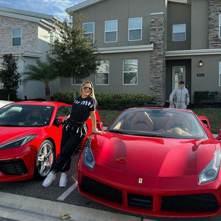 A imagem mostra uma mulher posando ao lado de dois carros esportivos vermelhos, um deles é uma Ferrari. Ela está vestindo roupas pretas e óculos de sol. Ao fundo, há uma casa com janelas grandes e uma pessoa ao lado da porta de entrada. Algumas árvores e arbustos completam a cena.