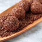 A imagem mostra brigadeiros sobre uma bandeja de madeira. Os doces estão cobertos com granulado de chocolate, dando uma textura característica. A bandeja está repleta de granulados, criando um visual harmônico. O fundo é claro, destacando ainda mais a cor marrom dos brigadeiros. A composição é simples e elegante, ressaltando a popularidade do doce em festas e eventos no Brasil.