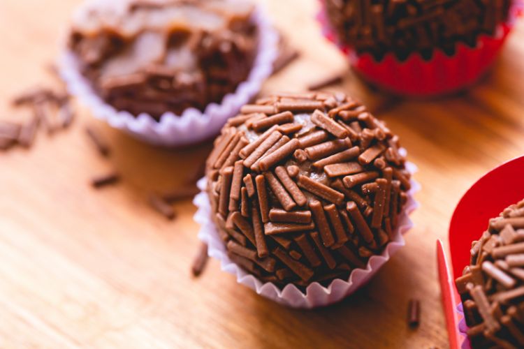 A imagem mostra brigadeiros, um doce tradicional brasileiro. Eles estão cobertos com granulado de chocolate e colocados em forminhas de papel coloridas. A textura do granulado dá um aspecto crocante ao doce. O fundo é de madeira, destacando a cor marrom dos brigadeiros. Há alguns granulados espalhados ao redor, adicionando um toque casual à apresentação. A composição é simples, mas atrativa, enfatizando a popularidade e o apelo visual desse doce em festas e celebrações.