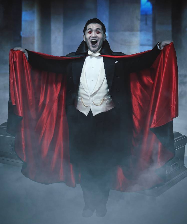 A imagem mostra um homem vestido como um vampiro clássico, com uma capa preta de forro vermelho, aberta dramaticamente. Ele usa um smoking elegante com colete branco e gravata borboleta. O cenário é nebuloso, evocando uma atmosfera misteriosa e sombria. Sua expressão é teatral e envolvente, capturando a essência de um personagem de filme de terror.