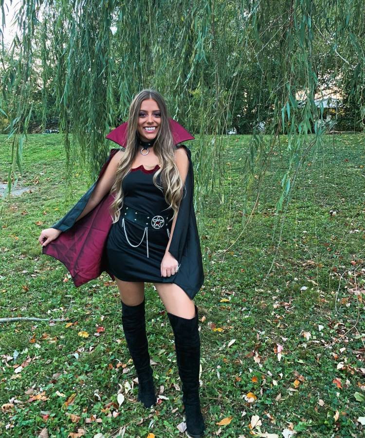 A imagem mostra uma mulher em um ambiente ao ar livre, vestida com uma fantasia de vampira. Ela usa um vestido preto curto, botas de cano alto e uma capa preta com forro vermelho. Um cinto com detalhes metálicos complementa o visual. Ela está sorrindo, cercada por grama verde e folhas caídas, em um cenário natural.