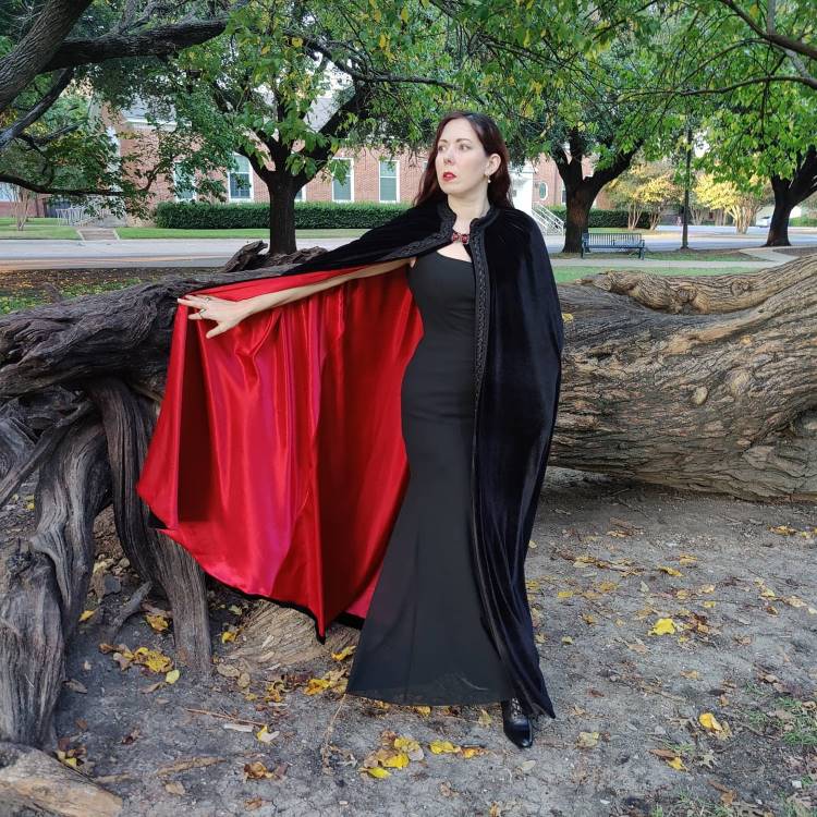 A imagem mostra uma mulher em um vestido preto elegante, destacando-se em um cenário arborizado. Ela usa uma capa de Halloween preta com forro vermelho vibrante, aberta dramaticamente. A expressão séria e o ambiente outonal, com folhas caídas, criam uma atmosfera misteriosa e encantadora, perfeita para uma celebração temática de Halloween.