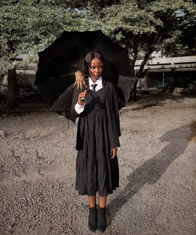 A imagem mostra uma mulher vestida como Wandinha Addams, ideal para fantasias femininas para o Halloween. Ela usa um vestido preto com gola branca e segura um guarda-chuva preto. O detalhe curioso é uma mão esquelética repousando sobre seu ombro, segurando o guarda-chuva com ela. O cenário externo, com árvores ao fundo, complementa o visual sombrio e misterioso, típico da personagem icônica da Família Addams.